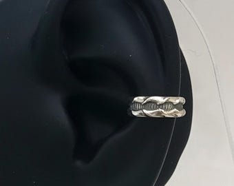 May include: A silver ear cuff with a woven design. The cuff is worn on a black ear model, showcasing the jewelry's intricate details and texture. The ear cuff is a stylish accessory.