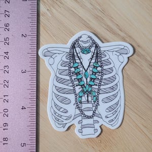 May include: A white sticker with a black outline of a ribcage and a turquoise and silver necklace with turquoise stones. The sticker is approximately 2 inches tall.