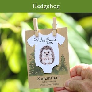 Puede incluir: Una tarjeta rectangular de papel kraft con un recorte blanco en forma de body, una ilustraci&oacute;n de un erizo y el texto Woodland BABY. Debajo, la tarjeta dice Samantha's BABY SHOWER WILDFLOWER SEEDS. Dos pinzas de madera sujetan el recorte.