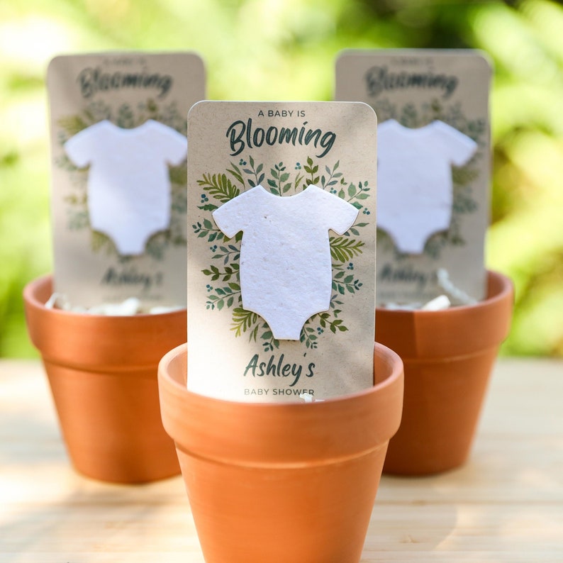 May include: Three terracotta pots each hold a baby shower favor. Each favor features a white onesie cutout on a card with the text "A Baby is Blooming" and "Ashley's Baby Shower". The cards are decorated with green foliage.