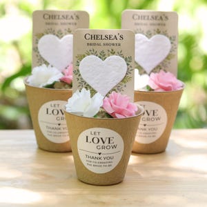 May include: Three small tan paper pots, each containing a white heart-shaped cutout and two artificial roses in white and pink. Each pot features a label with the text Chelsea's Bridal Shower and Let Love Grow Thank You For Celebrating The Bride-To-Be.