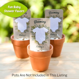 May include: Three terracotta pots each hold a baby shower favor. Each favor features a white onesie shape on a card with the text "A Baby is Blooming" and "Ashley's Baby Shower". The top left corner has a green leaf with the text "Fun Baby Shower Favors".