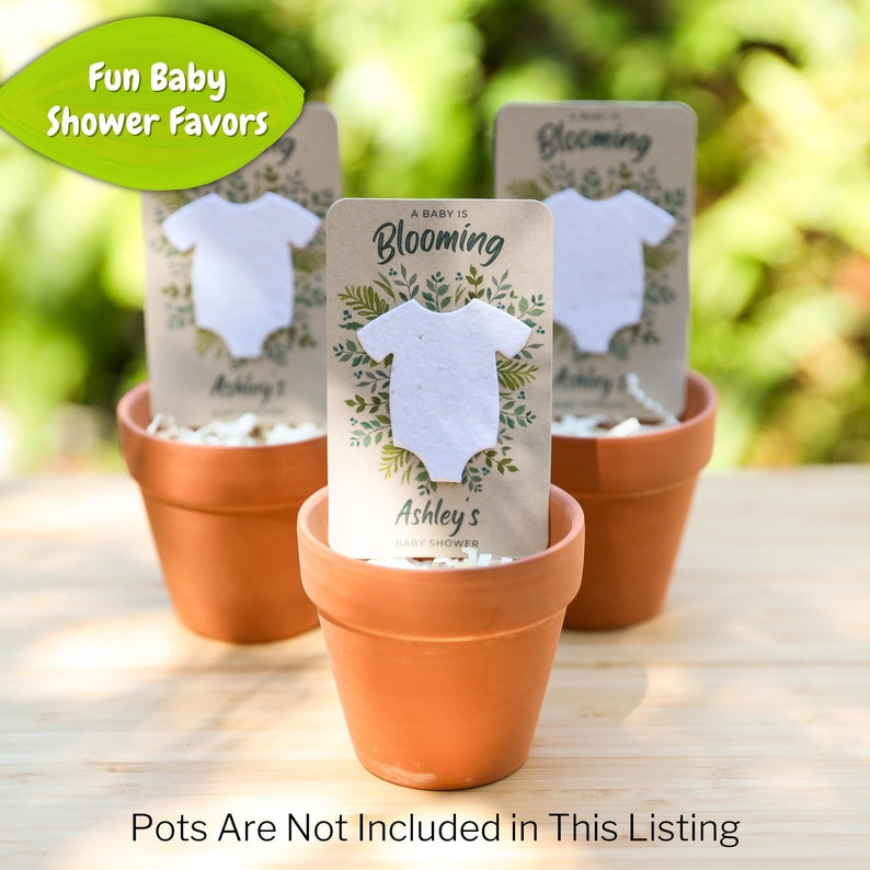 May include: Three terracotta pots each hold a baby shower favor. Each favor features a white onesie shape on a card with the text "A Baby is Blooming" and "Ashley's Baby Shower". The top left corner has a green leaf with the text "Fun Baby Shower Favors".