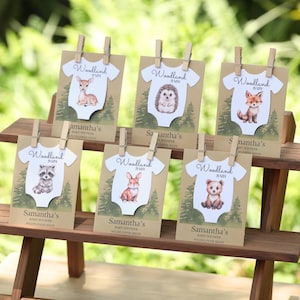 Puede incluir: Seis paquetes de semillas rectangulares en un soporte de madera, cada uno con un recorte de body blanco con una ilustraci&oacute;n de animal del bosque, el texto Woodland Baby y Samantha's Baby Shower Wildflower Seeds, sujetos por una pinza de madera.