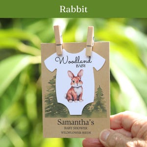 Puede incluir: Una tarjeta rectangular de papel kraft con un recorte en forma de body blanco, una ilustraci&oacute;n de un conejo marr&oacute;n y el texto Woodland BABY. Debajo, el texto dice Samantha's BABY SHOWER WILDFLOWER SEEDS. Dos clips de madera sujetan la tarjeta.