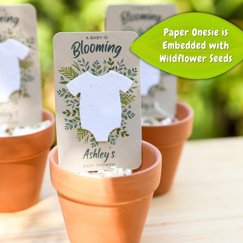 May include: Three terracotta pots each hold a baby shower favor. Each favor includes a paper onesie embedded with wildflower seeds, set against a card that reads "A Baby is Blooming" and "Ashley's Baby Shower."