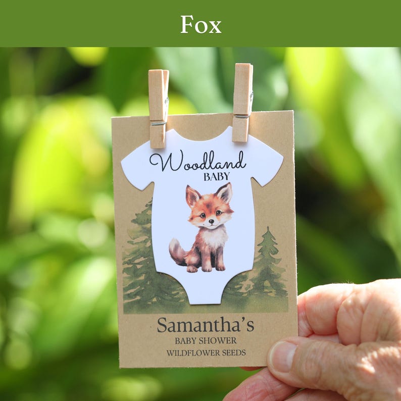 Puede incluir: Una tarjeta rectangular de papel kraft con un recorte de body blanco, una ilustraci&oacute;n de un zorro y el texto Woodland BABY. Debajo, la tarjeta dice Samantha's BABY SHOWER WILDFLOWER SEEDS. Dos pinzas de madera sujetan la parte superior.