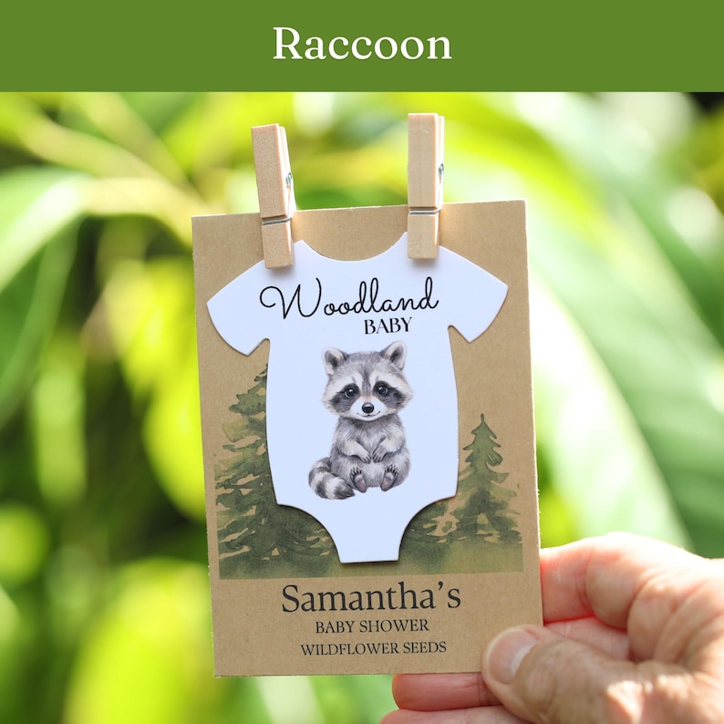 Puede incluir: Una tarjeta rectangular de papel kraft con un recorte blanco en forma de body, una ilustraci&oacute;n de un mapache y el texto Woodland BABY. La tarjeta incluye el texto Samantha's BABY SHOWER WILDFLOWER SEEDS y se sujeta con dos pinzas de madera.
