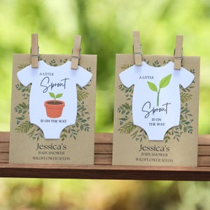 May include: Two rectangular kraft paper cards featuring a white onesie-shaped cutout attached with wooden clips. The cards display green botanical illustrations, the text A LITTLE SPROUT IS ON THE WAY, and Jessica's BABY SHOWER WILDFLOWER SEEDS.