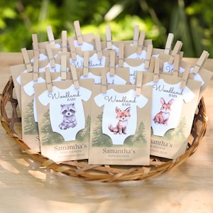 Puede incluir: Una colecci&oacute;n de sobres de semillas de papel kraft con forma de bodies de beb&eacute;, cada uno sujeto con una pinza de madera. Los sobres presentan ilustraciones de un mapache, zorro o conejo, con el texto Woodland BABY y Samantha's BABY SHOWER WILDFLOWER SEEDS.