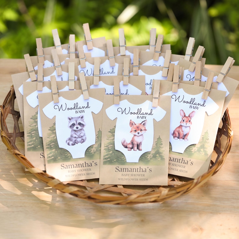Puede incluir: Una colecci&oacute;n de sobres de semillas de papel kraft con forma de bodies de beb&eacute;, cada uno sujeto con una pinza de madera. Los sobres presentan ilustraciones de un mapache, zorro o conejo, con el texto Woodland BABY y Samantha's BABY SHOWER WILDFLOWER SEEDS.