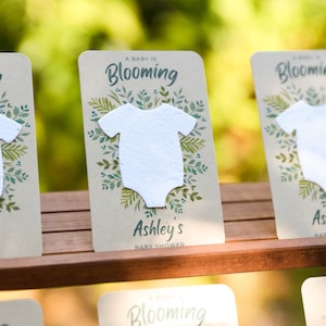 May include: Baby shower invitation cards with a white onesie cutout. The cards have the text "A Baby is Blooming" and "Ashley's Baby Shower" with a watercolor floral design. The cards are on a wooden surface.