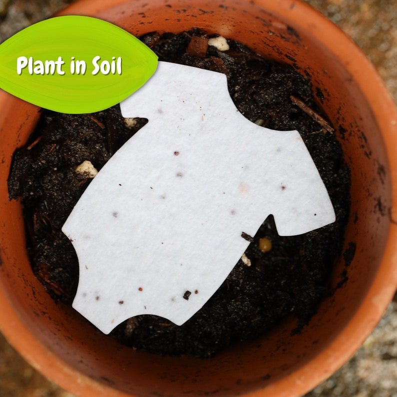 May include: A white, baby onesie-shaped seed paper in a terracotta pot filled with soil. The paper is dotted with seeds and is ready to be planted. A green leaf-shaped graphic with the text "Plant in Soil" is in the upper left corner.