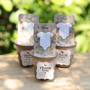 May include: Three small pots with baby-themed favors. Each pot has a card with a white onesie cutout and floral background, labeled "Baby Olivia." Attached tags read "Thank You for celebrating with us."
