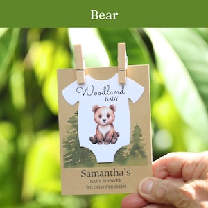 Puede incluir: Una tarjeta rectangular de papel kraft con un recorte blanco en forma de body con una ilustraci&oacute;n de un oso pardo, el texto Woodland BABY y el nombre Samantha's BABY SHOWER WILDFLOWER SEEDS, sujeta por dos peque&ntilde;as pinzas de madera sobre fondo verde.