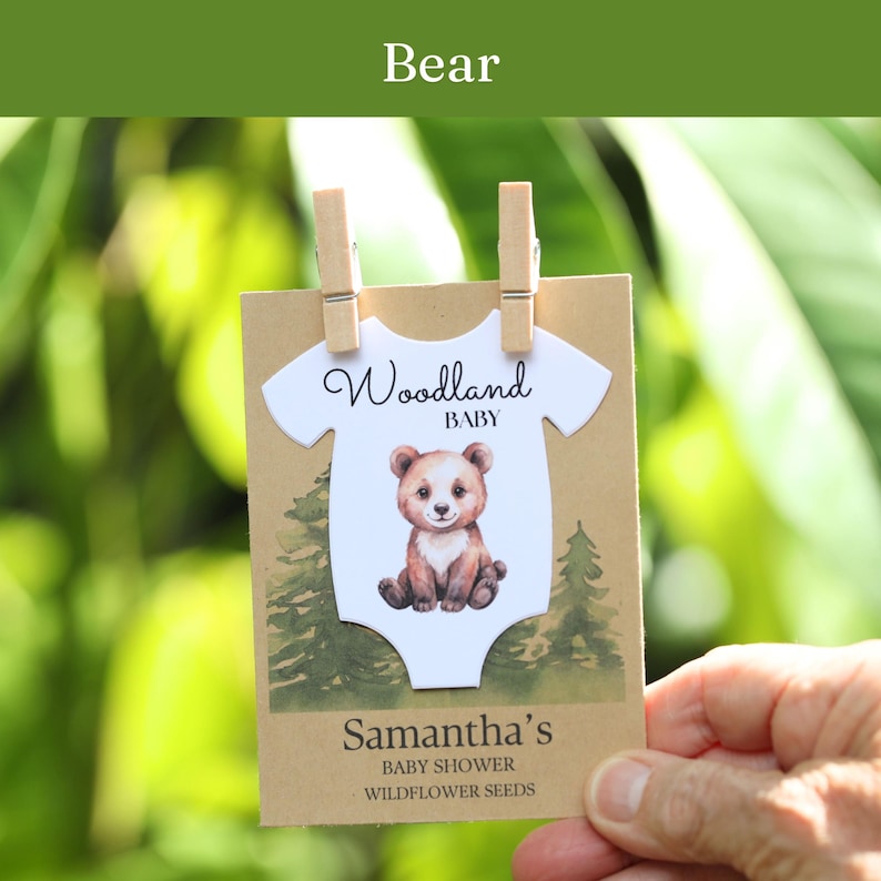 Puede incluir: Una tarjeta rectangular de papel kraft con un recorte blanco en forma de body con una ilustraci&oacute;n de un oso pardo, el texto Woodland BABY y el nombre Samantha's BABY SHOWER WILDFLOWER SEEDS, sujeta por dos peque&ntilde;as pinzas de madera sobre fondo verde.