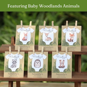 Puede incluir: Seis tarjetas rectangulares con fondos de papel kraft e ilustraciones de bosque verde, cada una con un recorte de body blanco con un animal del bosque en acuarela, sujetas con clips de madera. Texto Woodland Baby, Samantha's Baby Shower Wildflower Seeds.