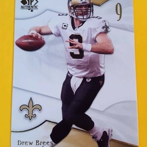 Puede incluir: Una tarjeta de fútbol blanca y dorada con Drew Brees, mariscal de campo de los New Orleans Saints. La tarjeta tiene un fondo dorado con un borde blanco y un logotipo de flor de lis dorado. La tarjeta también presenta el logotipo de SP Authentic y el número 9.