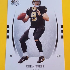 May include: A white and silver football card featuring a photo of Drew Brees, a professional football player, wearing a black and gold uniform with the number 9. The card has the text "SP Authentic" and "Drew Brees Saints" printed on it.