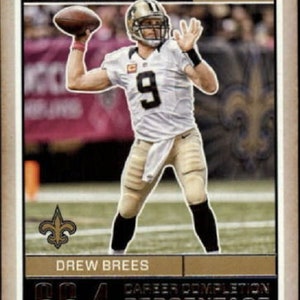 Pode incluir: Um cartão de futebol dourado e preto com Drew Brees, um quarterback dos New Orleans Saints. O cartão tem um fundo dourado com uma borda preta e apresenta uma foto de Brees lançando uma bola de futebol. O cartão também inclui o texto "Record Breakers" e "Classics 2018". O cartão também inclui o texto "Porcentagem de conclusão de carreira" e "Recorde da NFL" com o número "66,4".