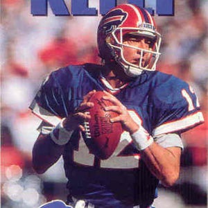 May include: A football player wearing a blue and red jersey with the number 12 on it, a blue helmet with a red stripe, and a white face mask. The player is holding a football in his right hand and is looking to the left. The word "KELLY" is written in blue letters above the player's head. The logo for SkyBox is in the bottom right corner of the image.
