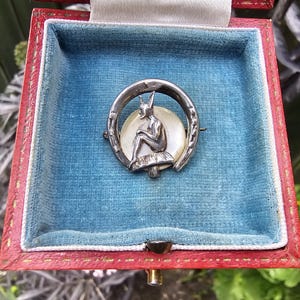 May include: A silver brooch featuring a seated figure on a mushroom, framed by a horseshoe shape, set against a circular mother-of-pearl inlay. The brooch is displayed inside a square red box with a light blue velvet interior lining.