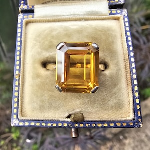 May include: A gold ring featuring a large, rectangular, emerald-cut yellow citrine gemstone held by four silver-toned prongs. The ring is displayed inside a square, navy blue velvet-lined jewelry box with gold-colored trim along the edges.