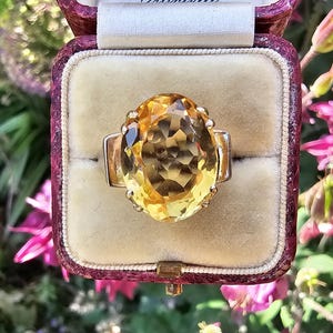 May include: A gold ring with a large, oval-cut yellow gemstone, set in a vintage jewelry box. The ring's design features a prominent central stone and a simple band. The box is burgundy with a cream-colored interior.