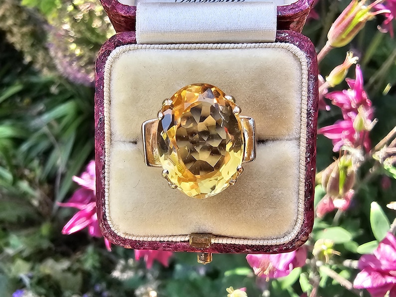 May include: A gold ring with a large, oval-cut yellow gemstone, set in a vintage jewelry box. The ring's design features a prominent central stone and a simple band. The box is burgundy with a cream-colored interior.