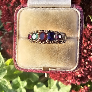 Puede incluir: Un anillo de oro con una serie de piedras preciosas de colores, incluyendo piedras rojas, verdes y azules. El anillo se muestra en un joyero forrado de terciopelo con exterior rojo. El fondo presenta flores rojas y follaje verde.