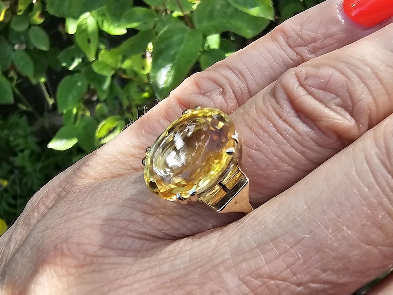 May include: A gold ring featuring a large, oval-cut yellow gemstone. The ring's setting has intricate details, and the gemstone is the focal point. The ring is on a person's finger, with a background of green foliage.