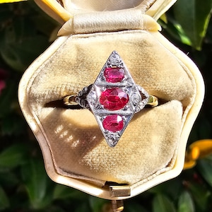 Puede incluir: Un anillo vintage con tres piedras de rubí engastadas en un diseño en forma de diamante. El anillo está engastado en una banda de oro y está en una pequeña caja de terciopelo.