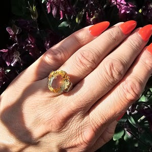 May include: A large, oval-shaped ring with a yellow-orange gemstone is displayed on a hand with bright orange nail polish. The ring is set in a silver-colored metal. The background features purple flowers and green foliage.