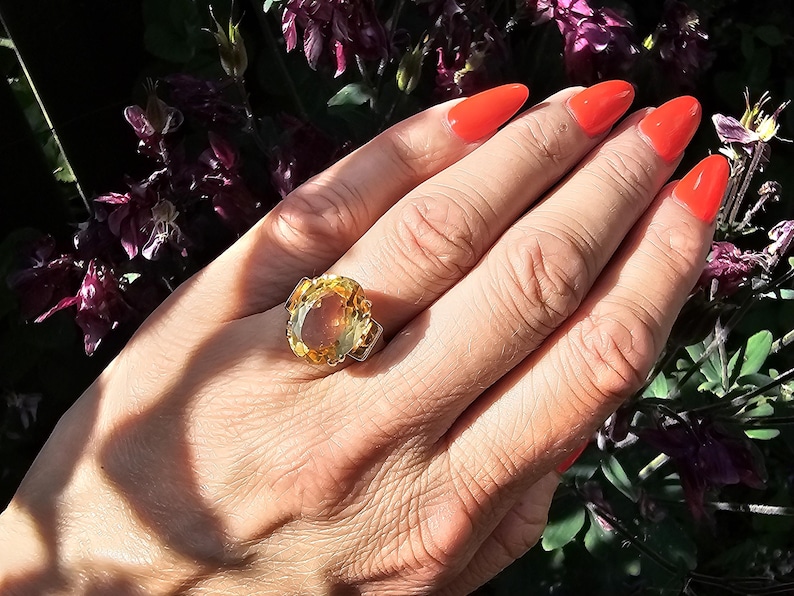 May include: A large, oval-shaped ring with a yellow-orange gemstone is displayed on a hand with bright orange nail polish. The ring is set in a silver-colored metal. The background features purple flowers and green foliage.