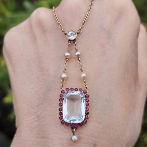 Puede incluir: Un collar con colgante dorado con una gran gema rectangular azul claro rodeada de pequeñas gemas rojas. El collar incluye perlas y una gema redonda y transparente en la parte superior.