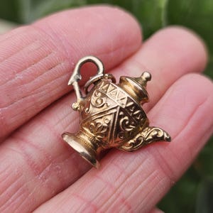 May include: A gold-colored teapot charm with intricate embossed designs. The small teapot has a curved spout, a handle, and a lid. The charm is held in a hand, with a blurred green background.
