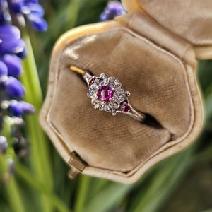 May include: A silver ring with a pink gemstone surrounded by smaller white gemstones. The ring is in a small, velvet-lined box.