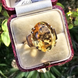 May include: A vintage gold ring with a large, oval-cut citrine gemstone. The ring is set in a gold band and is displayed in a burgundy velvet ring box. The gemstone has a warm, yellow-orange hue, and the ring is set against a cream-colored interior.
