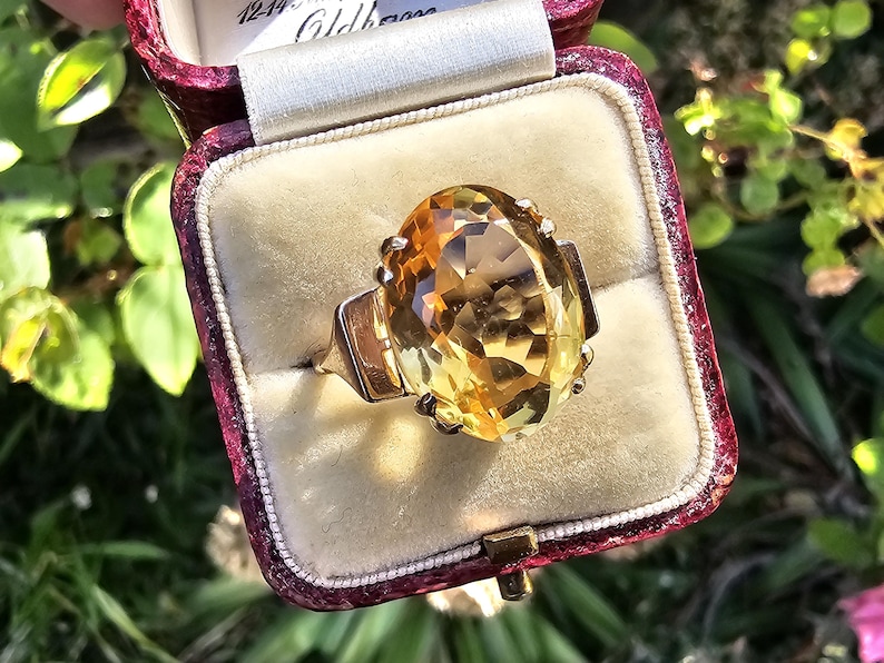 May include: A vintage gold ring with a large, oval-cut citrine gemstone. The ring is set in a gold band and is displayed in a burgundy velvet ring box. The gemstone has a warm, yellow-orange hue, and the ring is set against a cream-colored interior.