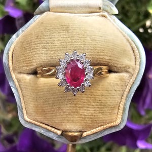 May include: A gold ring featuring a central oval-cut red ruby surrounded by a halo of small, clear, round-cut white diamonds. The band has a twisted design and is presented inside a beige velvet-lined jewelry box with a light blue trim.