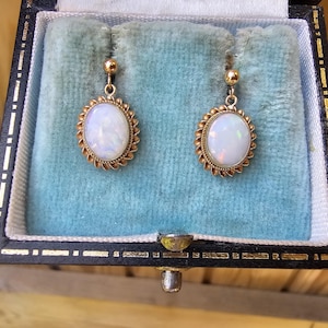 May include: A pair of gold drop earrings featuring oval-shaped opals surrounded by a twisted metal border. Each earring is suspended from a small gold ball stud. The earrings are displayed inside a black jewelry box with a light blue velvet lining.
