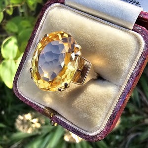 May include: A gold ring with a large, oval-cut yellow gemstone, set in a vintage-style mounting. The ring is displayed in a burgundy velvet-lined box. The gemstone has a faceted surface, catching the light. The ring is a piece of jewelry.