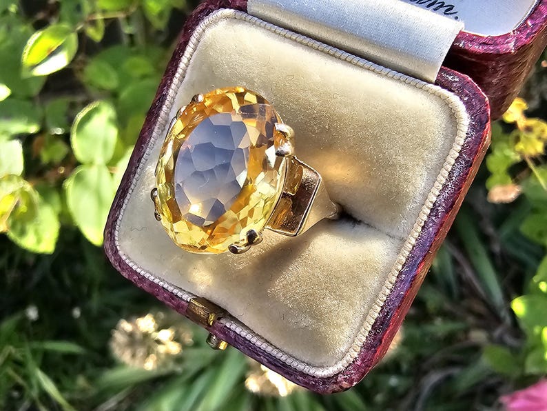 May include: A gold ring with a large, oval-cut yellow gemstone, set in a vintage-style mounting. The ring is displayed in a burgundy velvet-lined box. The gemstone has a faceted surface, catching the light. The ring is a piece of jewelry.