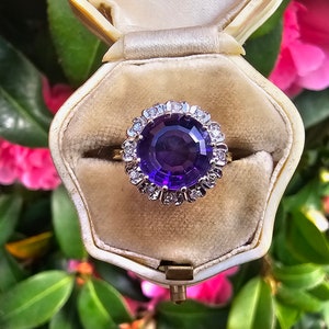 Puede incluir: Un anillo de amatista púrpura con un halo de pequeños diamantes engastados en oro amarillo. El anillo está en una caja de anillo blanca.