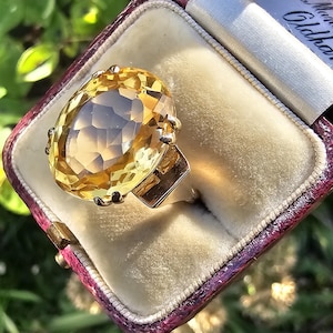 May include: A large, oval-cut citrine gemstone ring set in gold, displayed in a vintage maroon jewelry box with a cream-colored interior. The ring's facets catch the light, showcasing its warm yellow color. The box has text that reads "Manchester" and "Oldham."