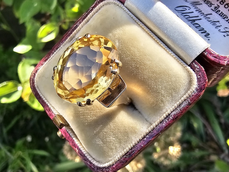 May include: A large, oval-cut citrine gemstone ring set in gold, displayed in a vintage maroon jewelry box with a cream-colored interior. The ring's facets catch the light, showcasing its warm yellow color. The box has text that reads "Manchester" and "Oldham."