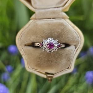 May include: A pink gemstone ring with a floral design set in a white metal band. The ring is in a white velvet ring box.
