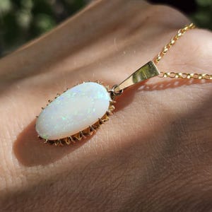 May include: An oval-shaped white opal pendant featuring iridescent flashes of blue and green light, set in a gold-toned prong setting. The pendant is attached to a delicate gold-toned chain, creating a classic necklace design with a polished finish.