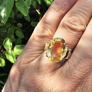 May include: A large, oval-cut yellow gemstone ring with a silver band. The gemstone has a gradient of yellow to orange hues. The ring is set with two rectangular accent stones on each side of the main stone.
