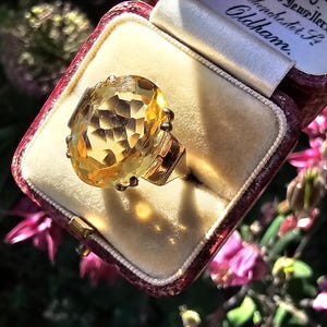 May include: A gold ring with a large, faceted, yellow gemstone, presented in a vintage maroon jewelry box with a cream-colored interior. The ring is the focal point, catching the light. The box is open, revealing the ring.